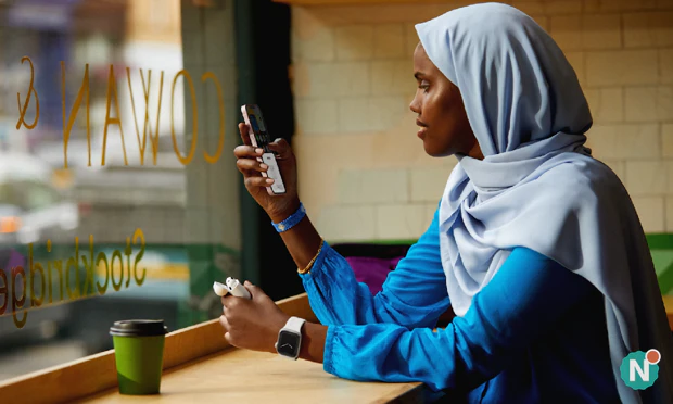 Vrouw in een koffietent kijk op haar iPhone terwijl ze de AirPods case vasthoudt en een Apple Watch draagt